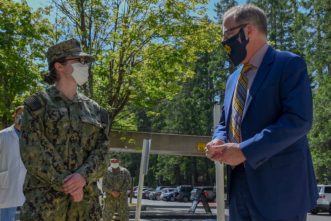 A man wearing a business suit speaks with a sailor outdoors. Both are wearing face masks.
