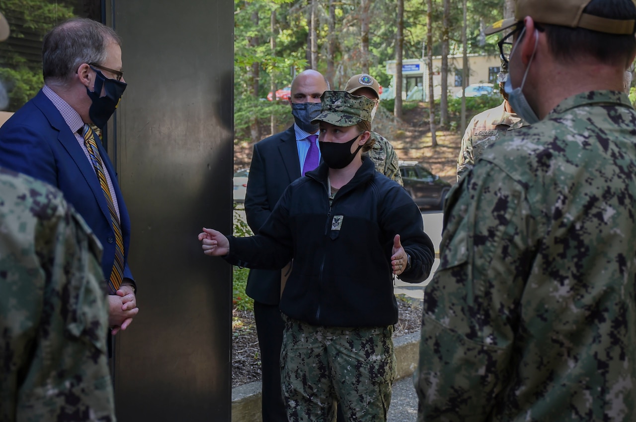 A man wearing a business suit speaks with a group of sailors outdoors. All are wearing face masks.