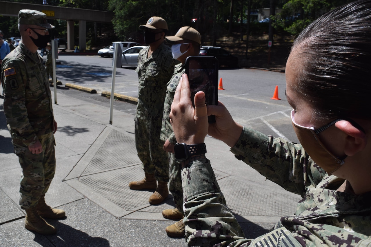 An Army general speaks outdoors with two sailors as another sailor takes their picture with her phone. All are wearing face masks.