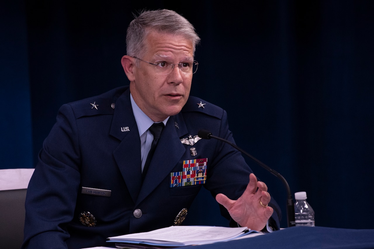 A man in military uniform sits at a table behind a microphone.