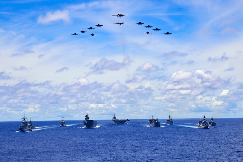 A triangular formation of aircraft flies above a formation of ships in blue sea.