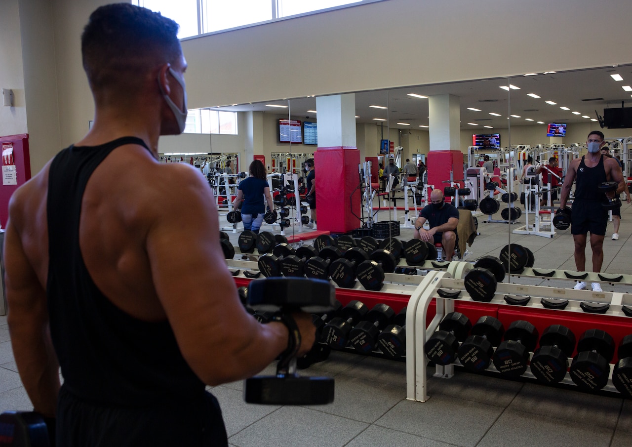 A man wearing a face mask watches his form in a mirror as he lifts dumbbells.