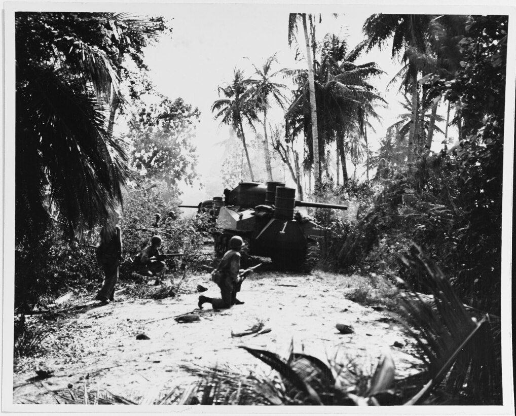 Men with rifles stand near a tank along a road in dense jungle.