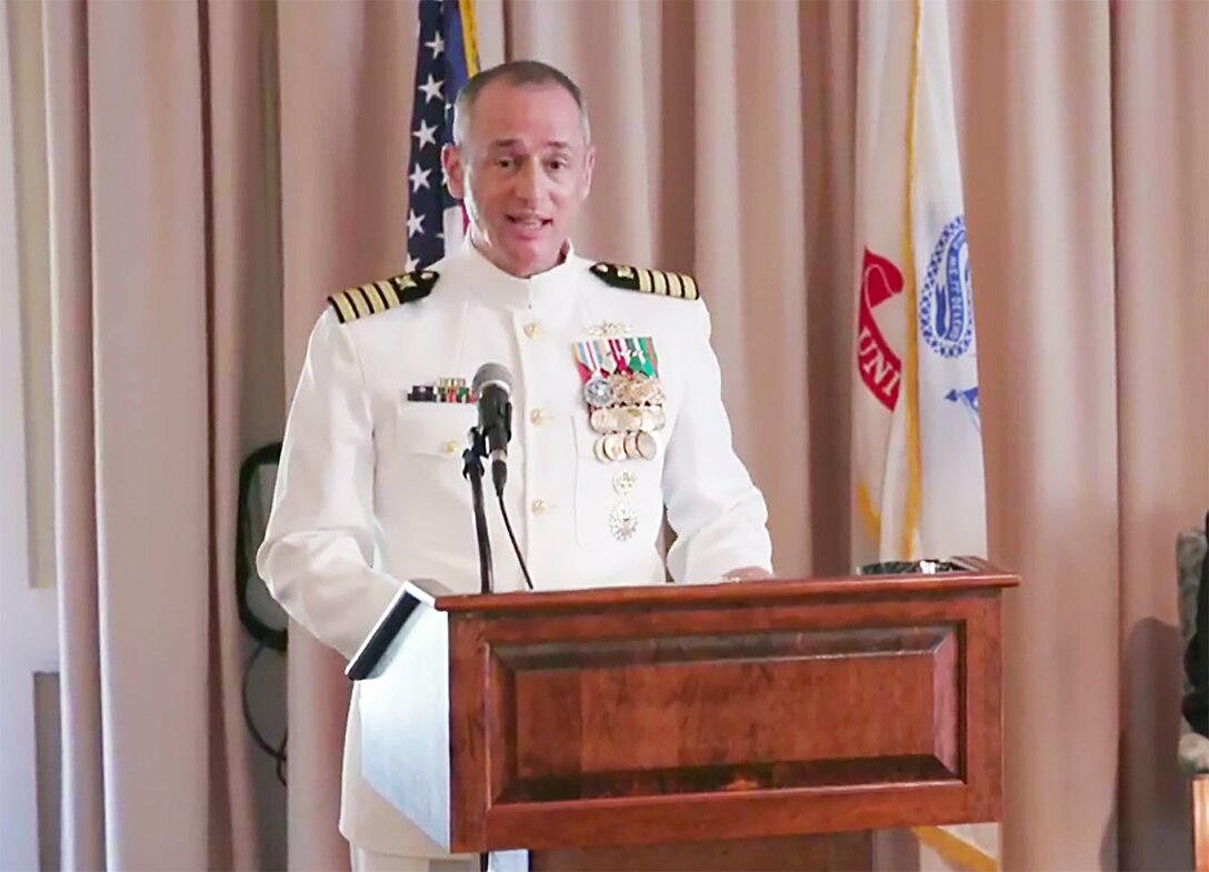 Navy Capt. Fred Dini stands behind a podium in his dress uniform as he speaks during a change of command ceremony.