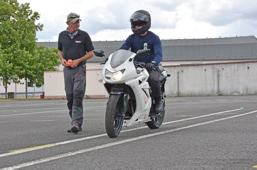 A soldier rides a motorcycle as an instructor stands next to him.