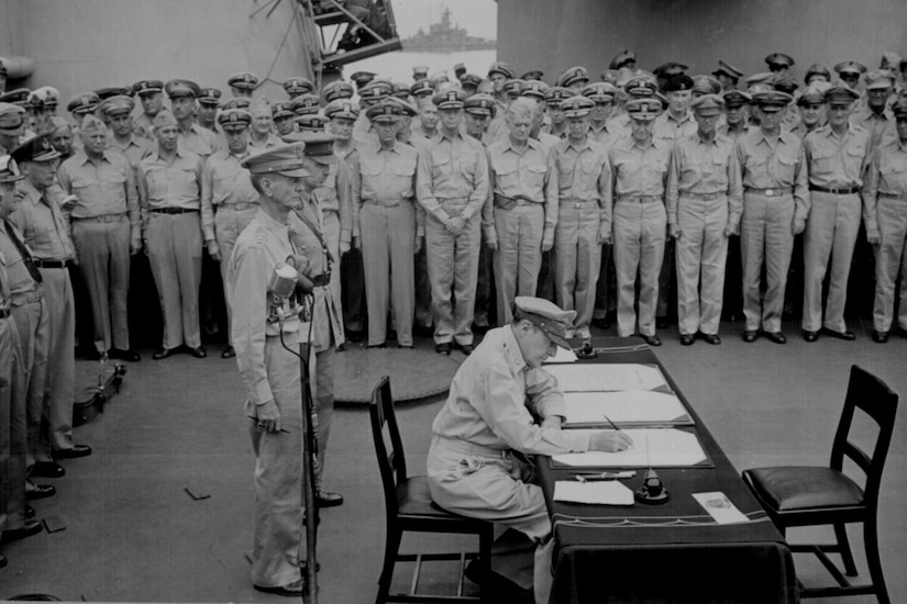 A military officer sits at a desk and signs papers while other officers stand around him.