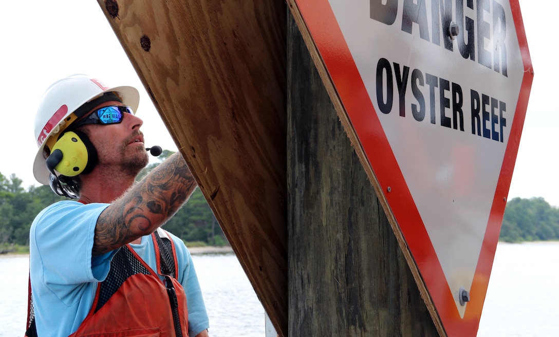 man in life jacket holds up sign that reads, "DANGER Oyster Reef" while placing it on a pole in the water.