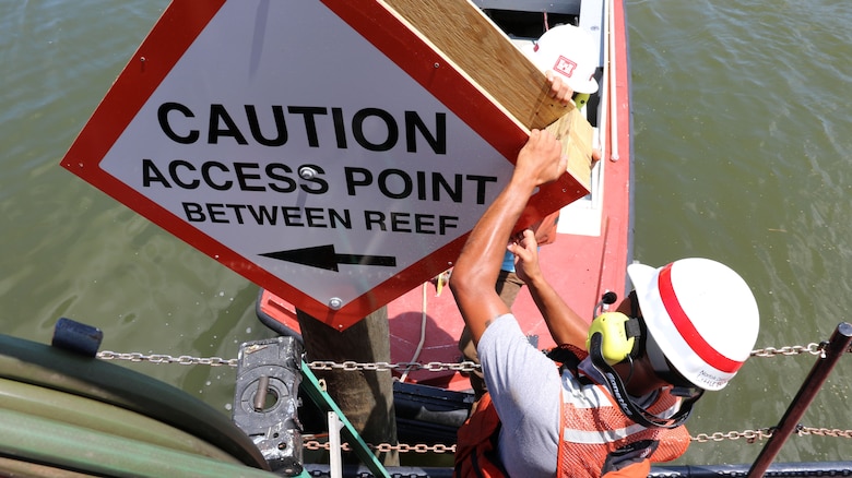 man in life jacket holds sign on a pole in the water that reads, "CAUTION Access Point Between Reefs"