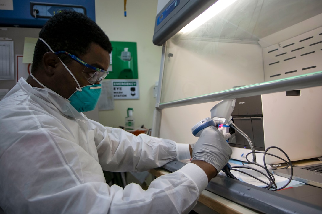 An Army lab technician wearing protective gear uses a scanner on an object he's holding under a lab hood.