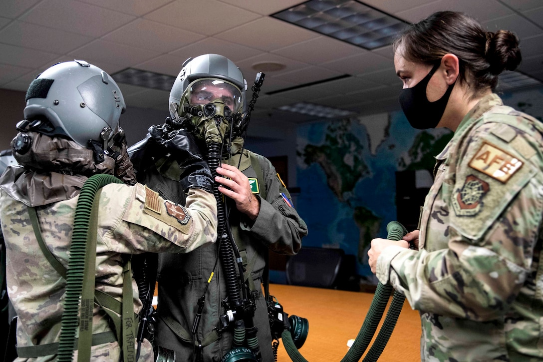 An airman in a helmet with a breathing apparatus adjusts the gear of an airman dressed similarly, as a third with a face mask observes.