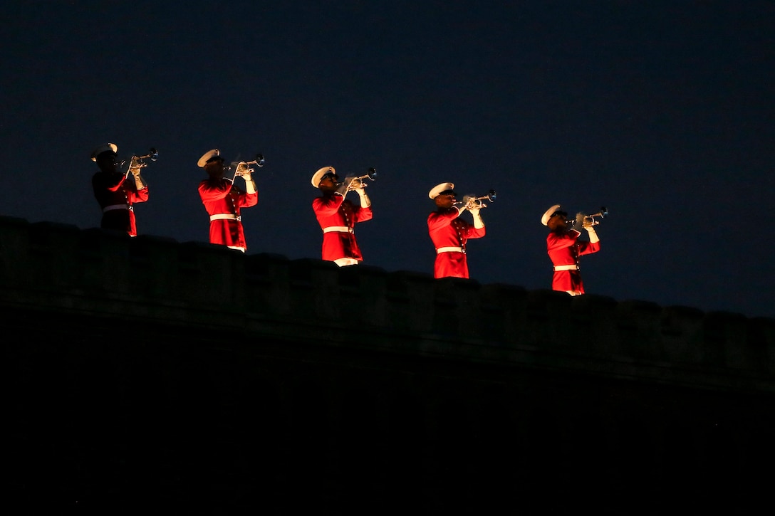 Five Marine Corps buglers perform in a line atop a building at night.