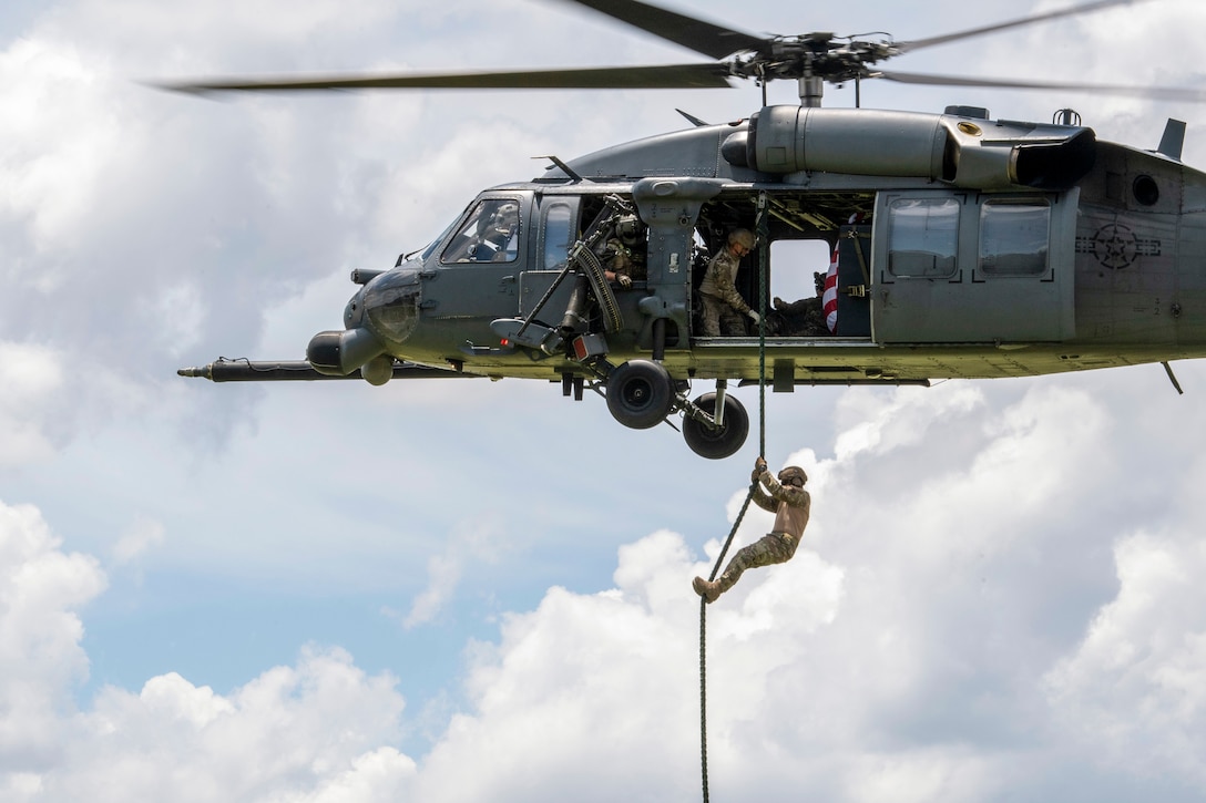 An airman fast-ropes out of a helicopter where others watch.