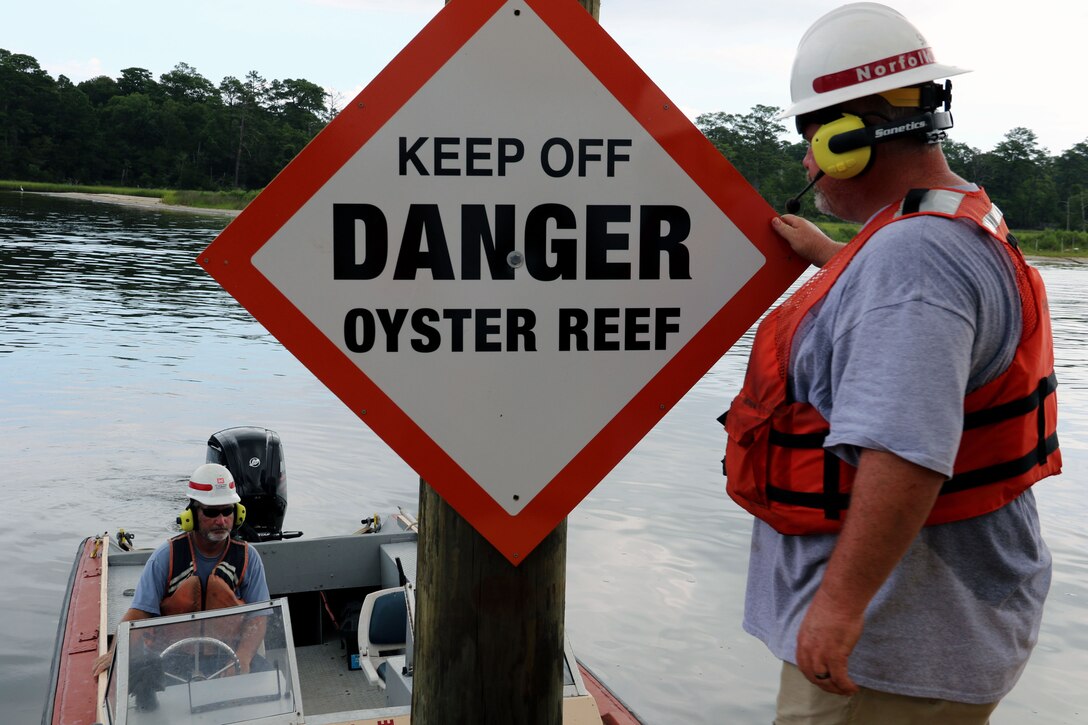 two men in life jackets, one in a small boat, are in the frame with a sign that says, "Keep Off Danger Oyster Reef"