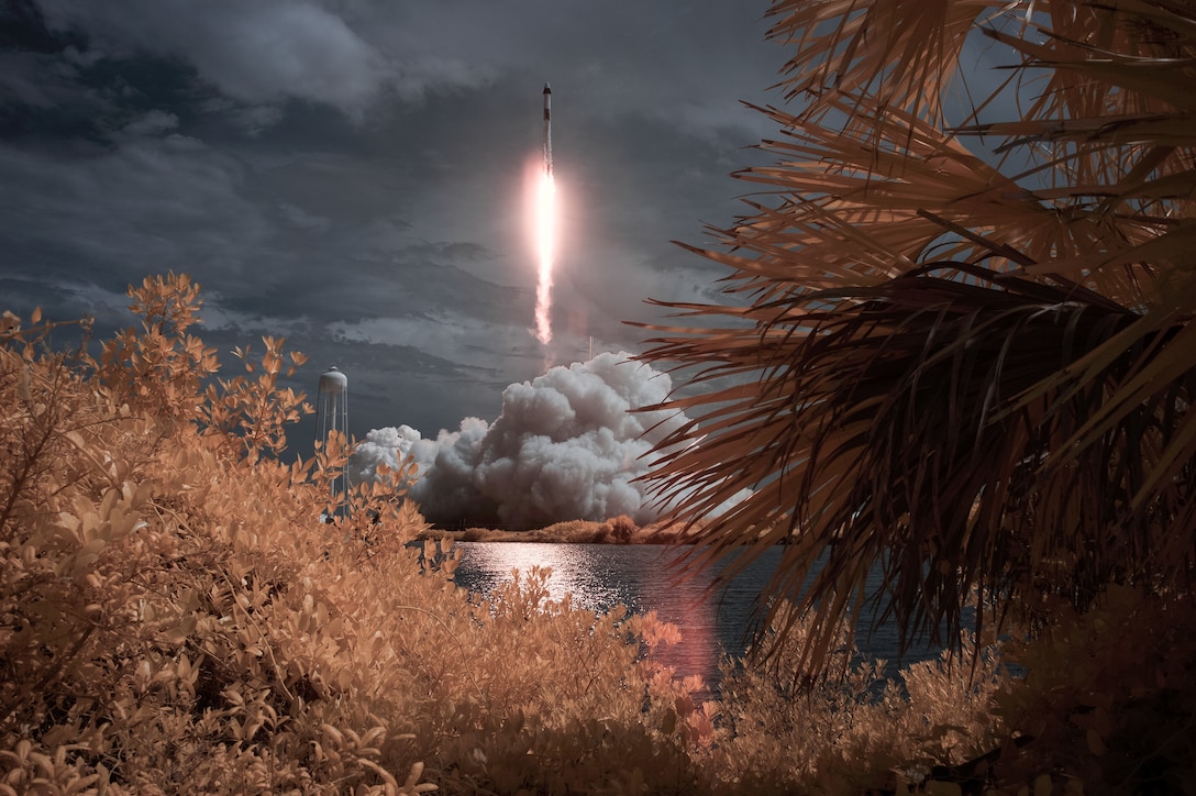 A flash of light and clouds of smoke are seen in the distance, framed by foliage, as a rocket takes off.