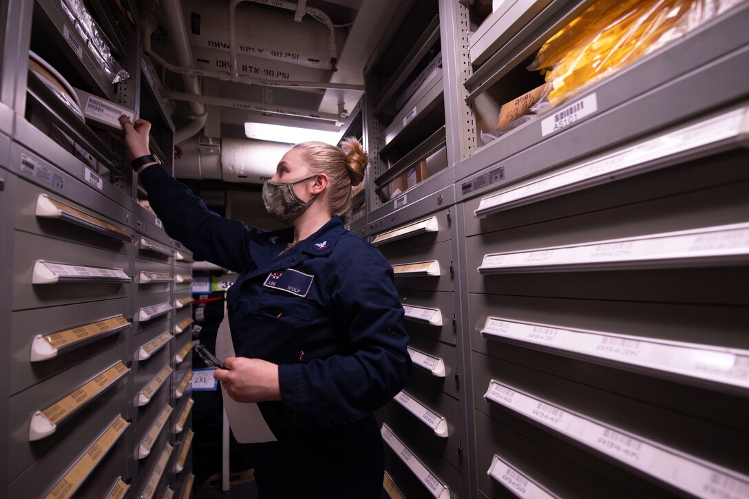A sailor puts items on one of several shelves in a small room.