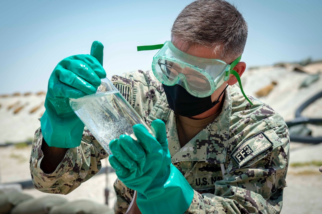 A sailor wearing a face mask, goggles and gloves holds a jar of liquid.