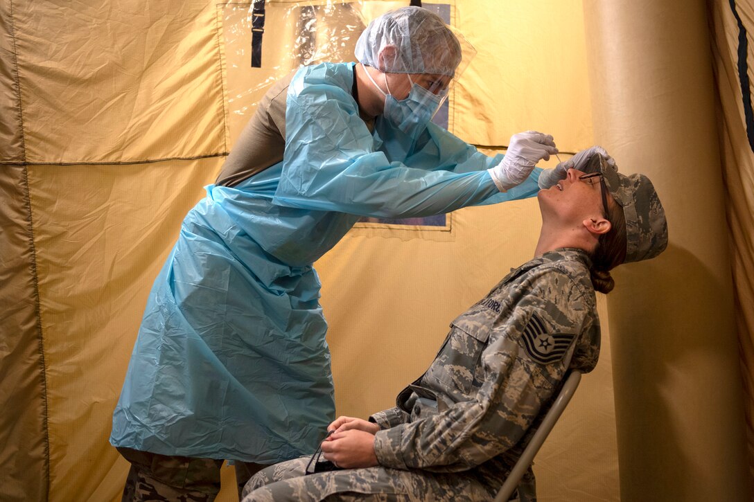 A standing soldier in protective gear swabs the nose of an airman sitting in a chair.