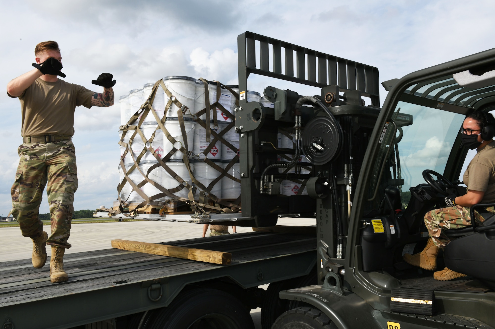 Photo shows one Airmen directing another on a forklift while she lowers a pallet containing numerous buckets to a flatbed truck.