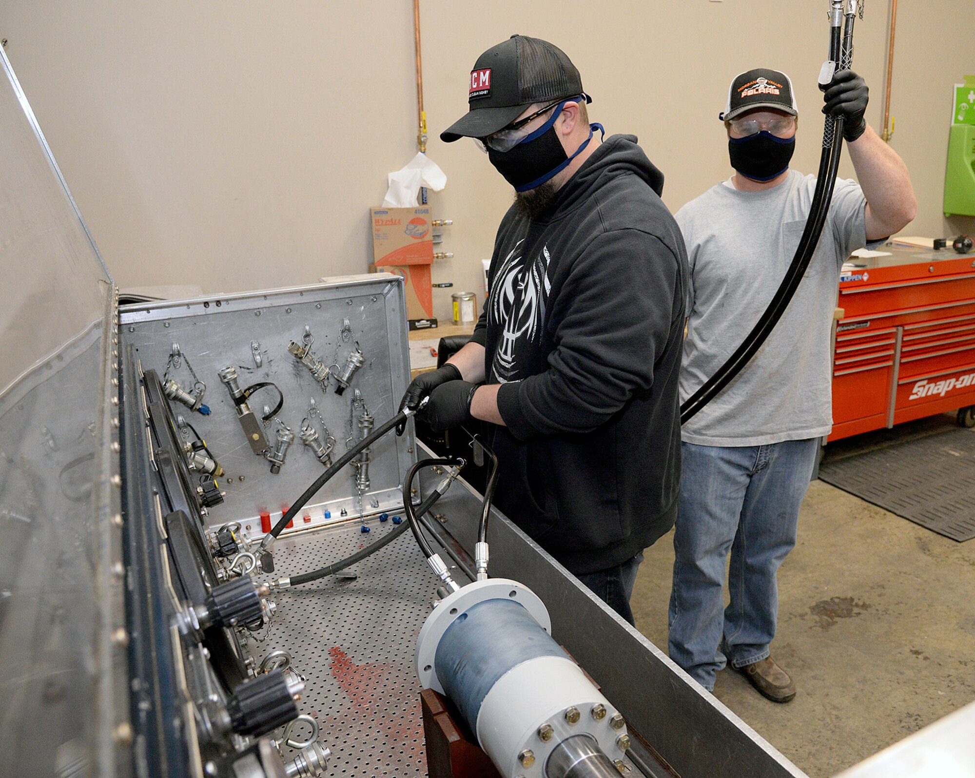 Shadd Straw and Garrett Kippen (left to right), 581 Missile Maintenance Squadron powered support system mechanics, wear their protective face coverings while they work to connect hydraulic lines from a test stand to a loading mechanism on Apr. 28, 2020, at Hill Air Force Base, Utah.