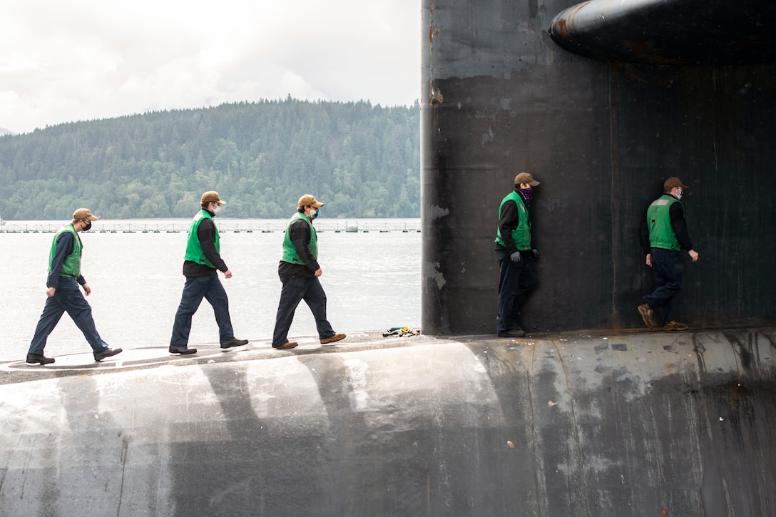 Sailors wearing green vests and protective gear walk in a line.