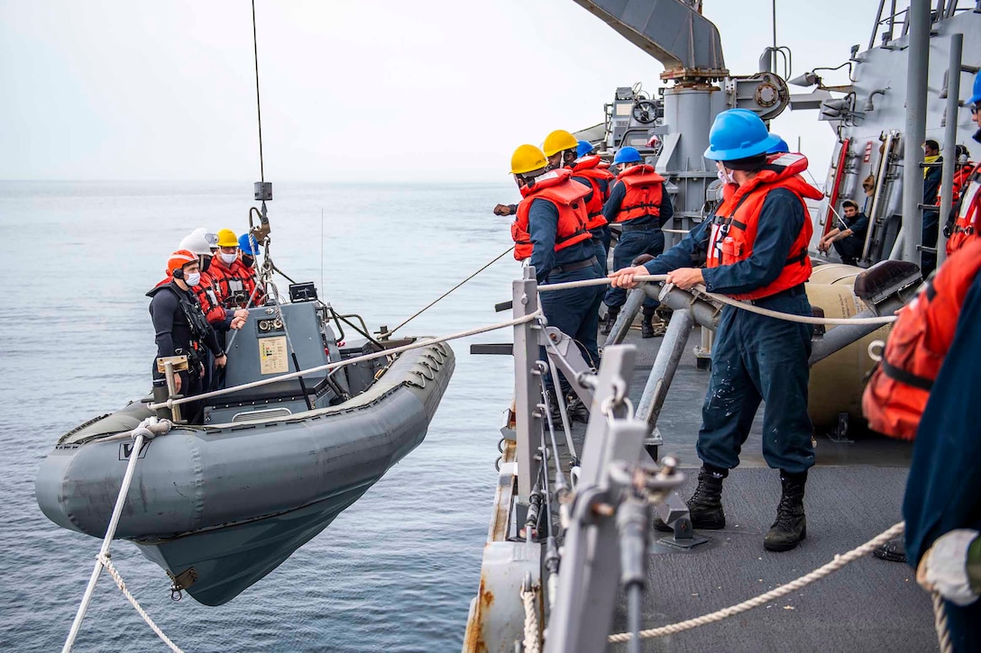 Sailors stand in an inflatable boat next to a ship as it lowers into water; other sailors stand on the ship watching.