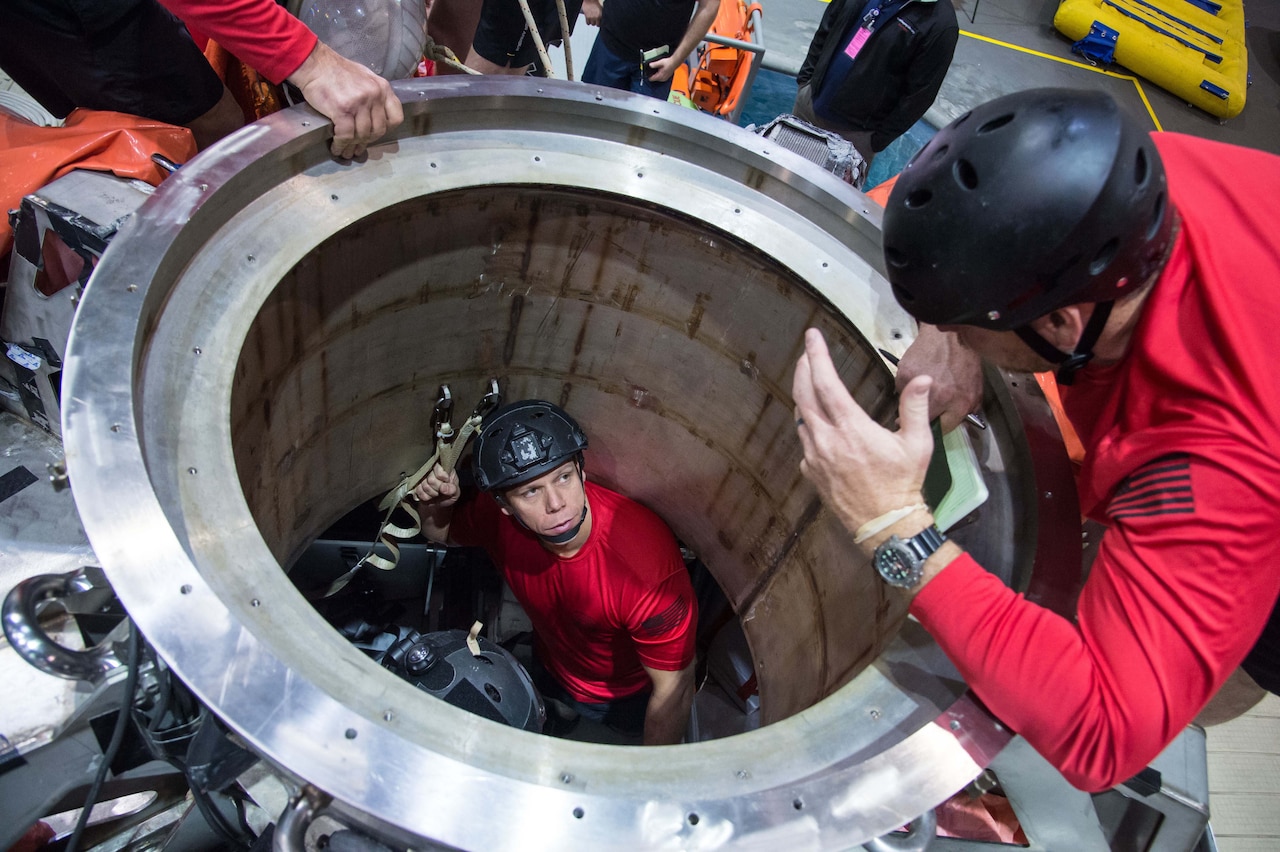 A young man in a red shirt with a helmet stands in the center of a metal ring.  Another individual in similar clothing looks down on him from above.