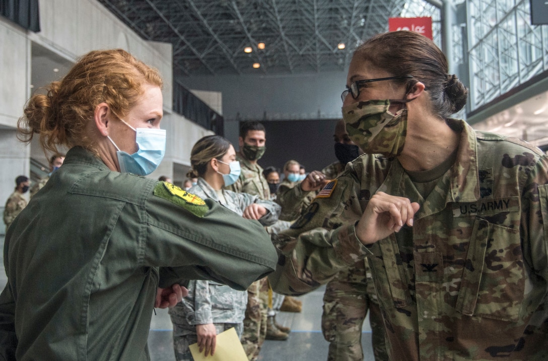 A soldier and an airman wearing face masks elbow-bump each other in a large room.