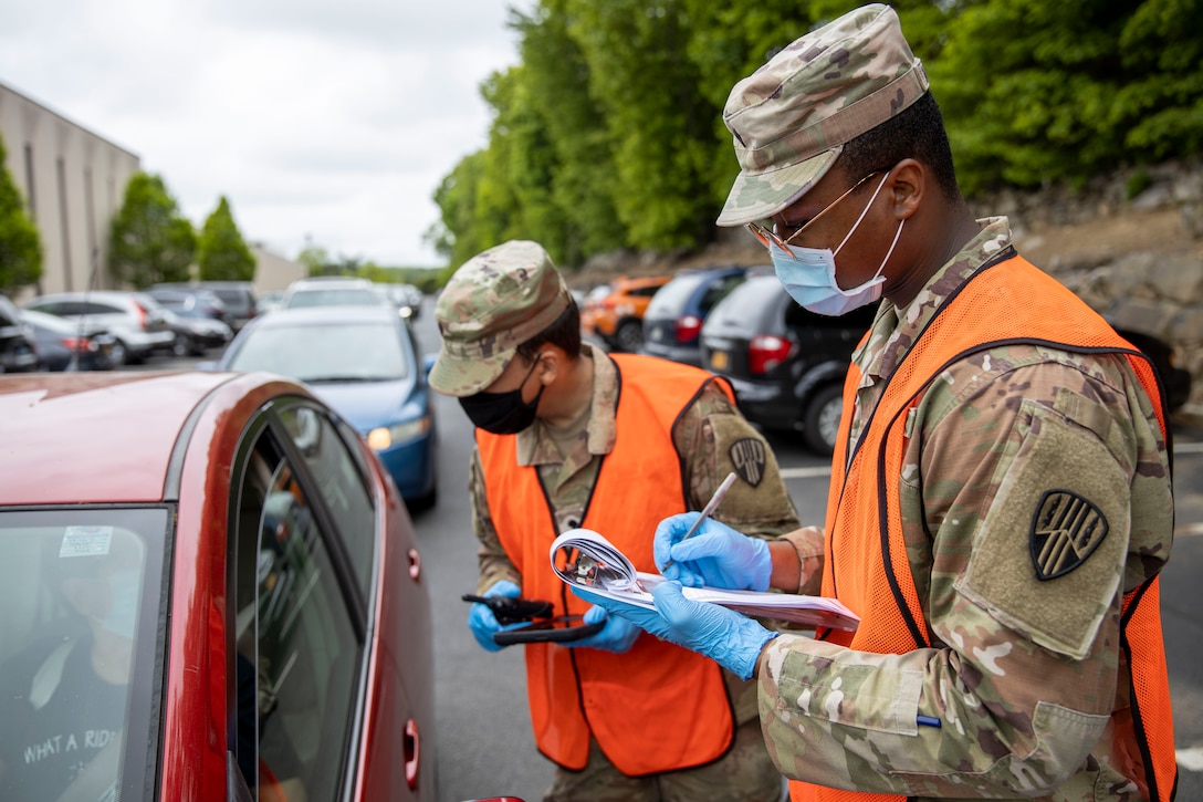 A soldier talks to a driver in a car while a second soldier takes notes.