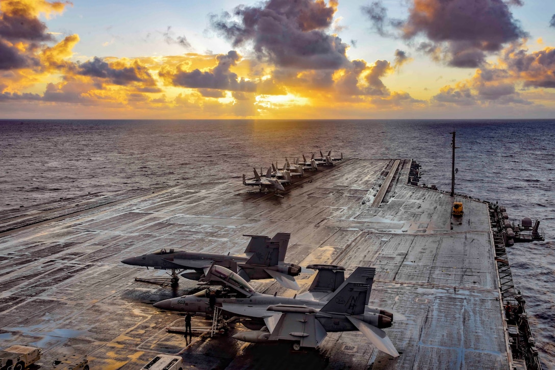An aircraft carrier with several military jets floats on the water at twilight.