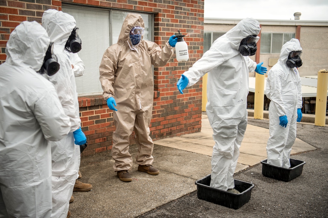 A civilian in protective gear sprays a service member in protective gear standing in a bin outside a building.