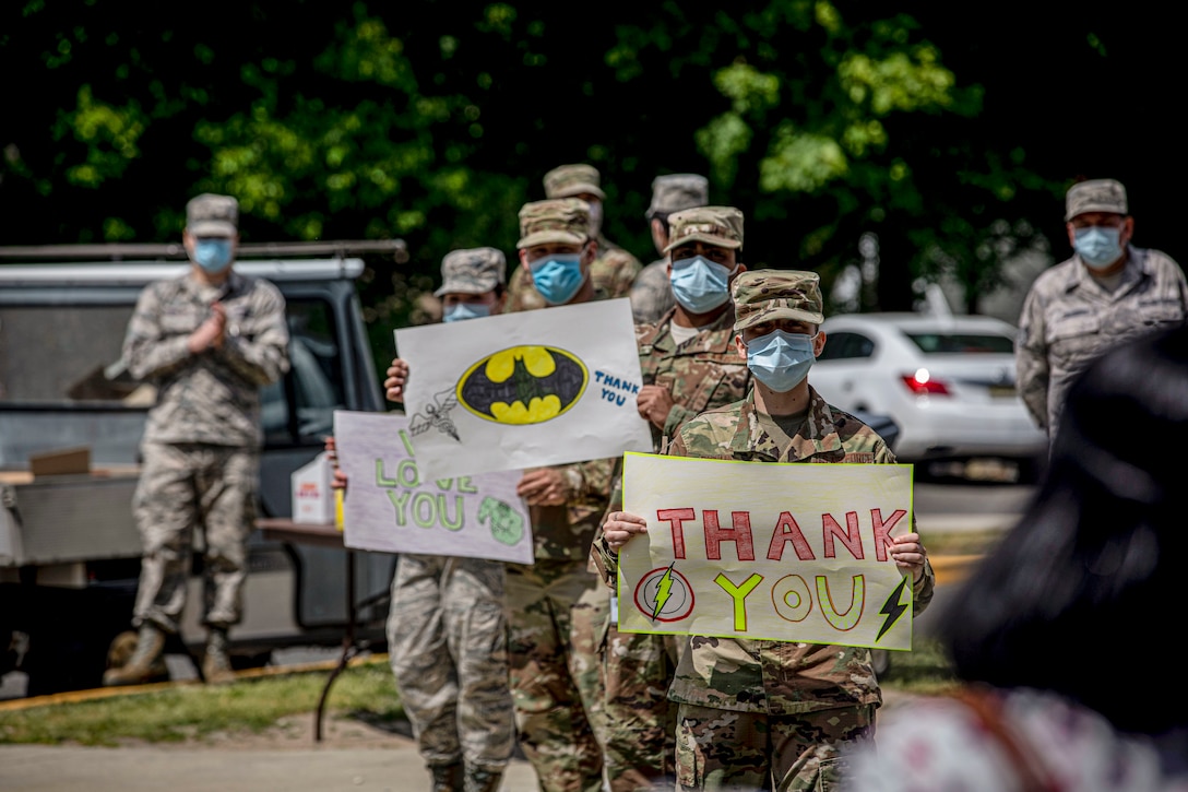 Airmen wearing masks hold up handwritten "Thank You" signs as a civilian walks toward them outside a building.