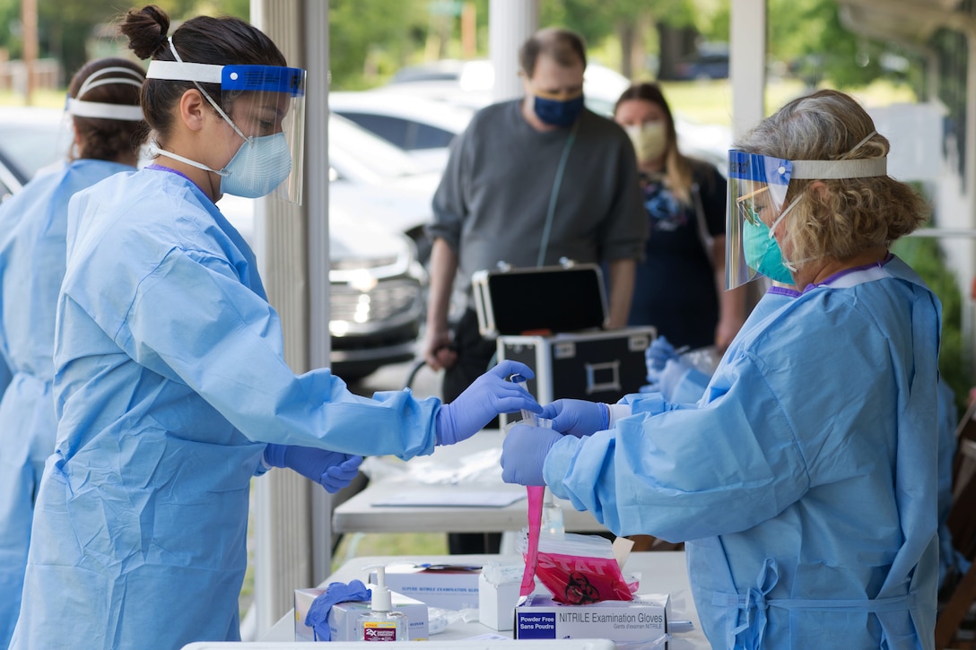 A soldier wearing personal protective equipment puts a COVID-19 test into a sterile bag while another soldier holds the plastic bag open for her.