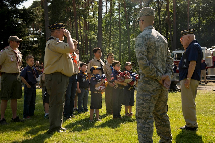 An airman stands by as a scout leader salutes an older veteran. Several young men stand with them, three of whom are holding unserviceable flags.