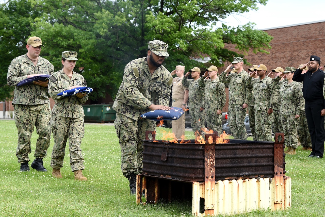 An airman places a folded flag into a fire pit as others nearby salute.