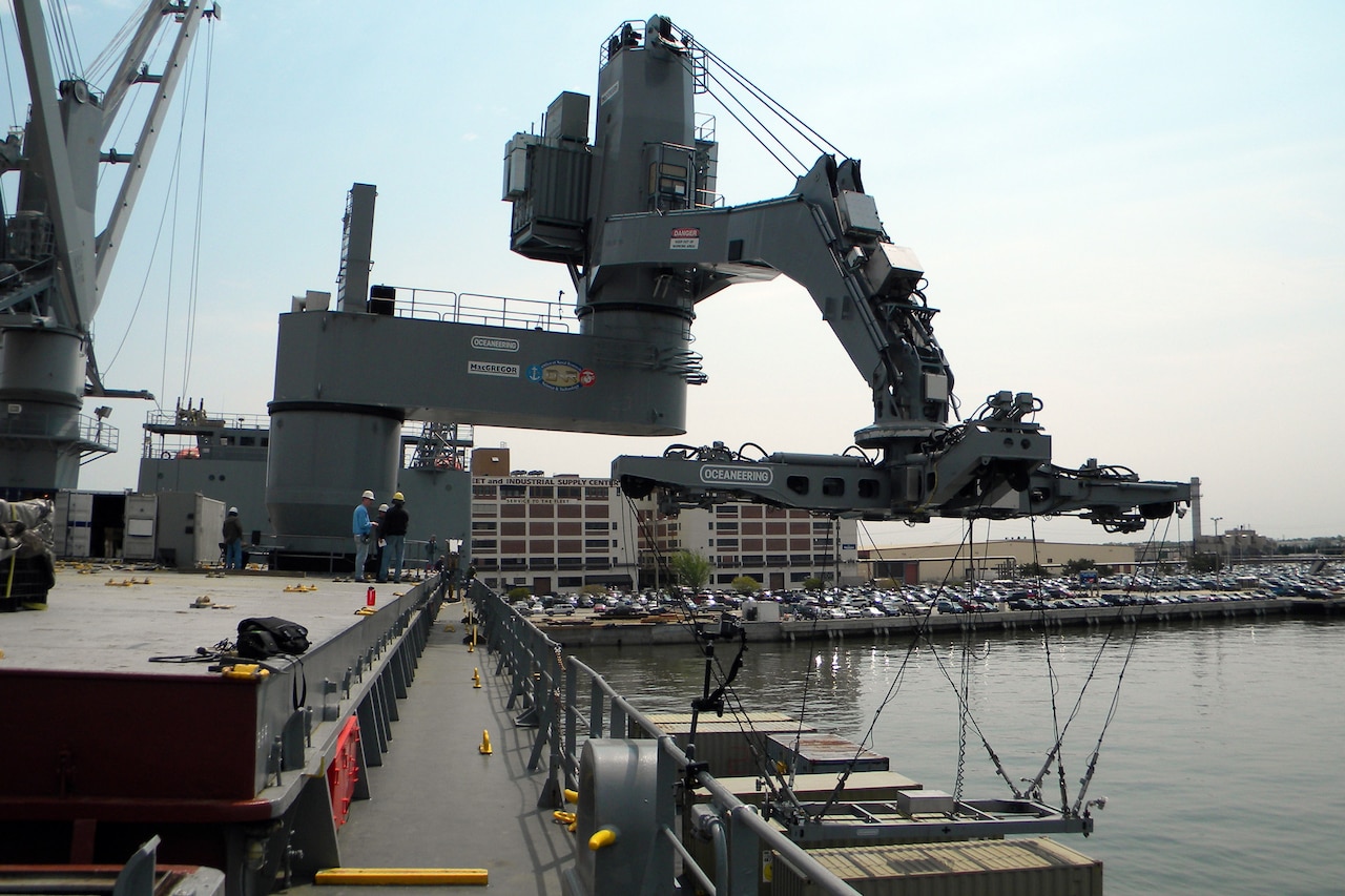 A crane, similar to a large robotic arm, moves toward a cargo container that is on a ship.