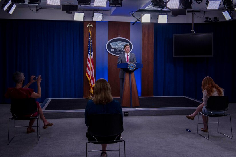 A man stands a lectern in front of three socially-distanced, seated women.