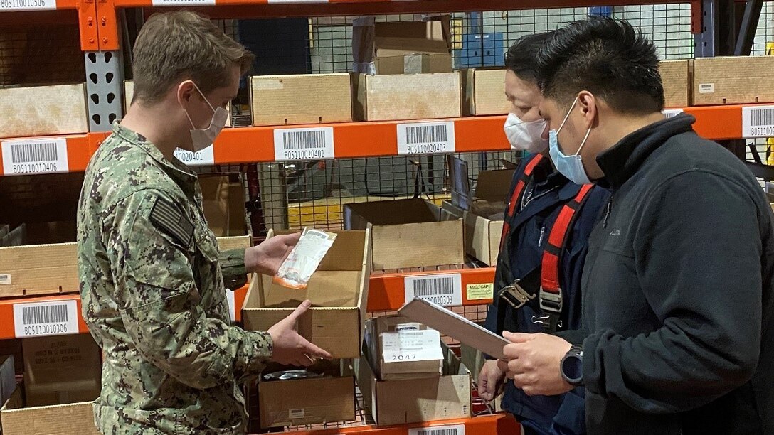 Three workers in face masks review materials pulled from a bin in front of a warehouse shelf lined with similar bins.