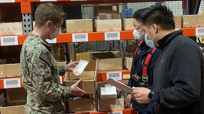 Three workers in face masks review materials pulled from a bin in front of a warehouse shelf lined with similar bins.