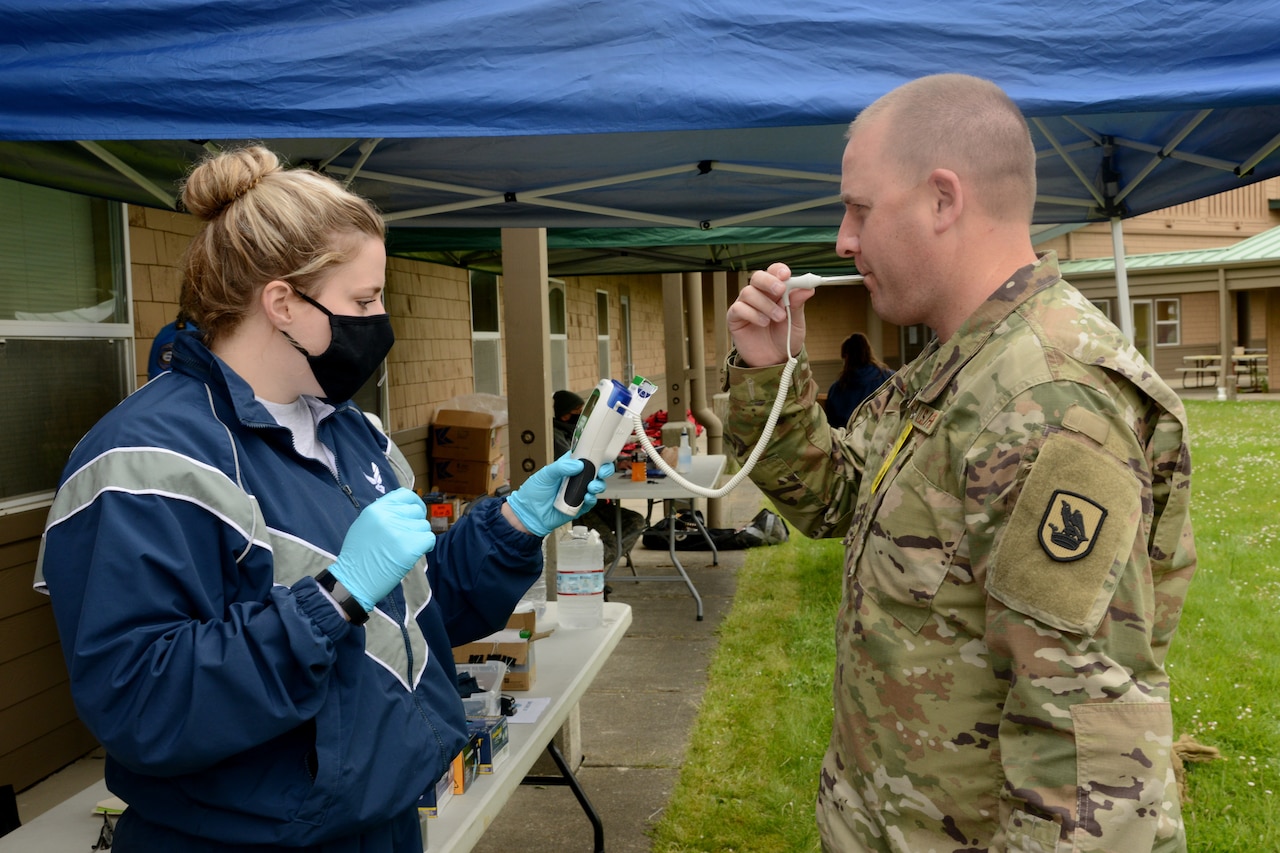 A woman wearing protective gear takes an airman's temperature.