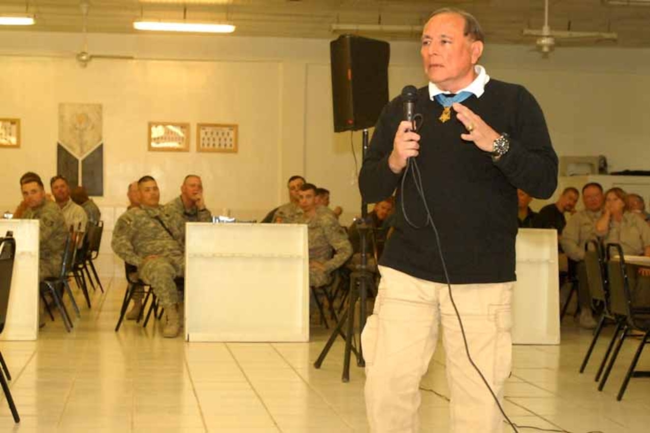 An older man with a microphone talks to several seated soldiers.