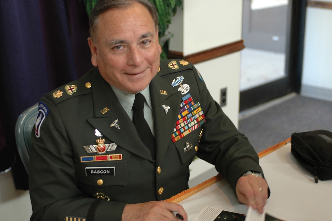 A man in a military uniform sits at a table autographing photos.