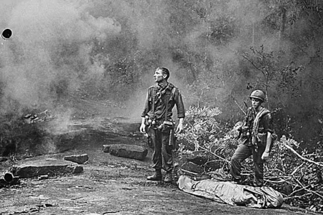 Two soldiers stand beside a body bag in the jungle.
