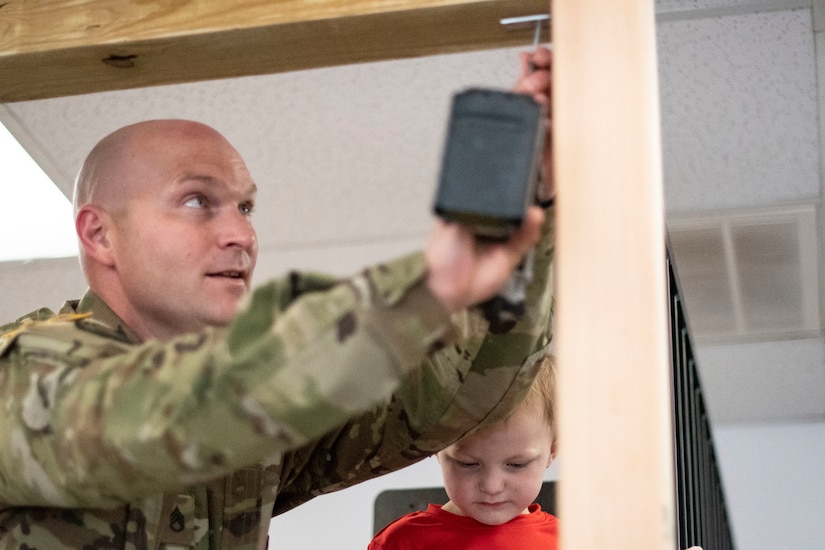 Man in green camouflage uniform holds drill as young boy in red t-shirt stands in the background.