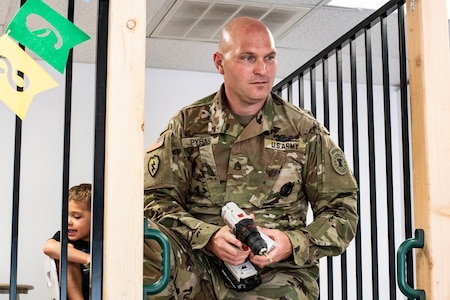 man in green camouflage uniform holds a drill while a young boy sits behind him.