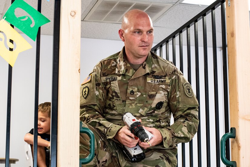 man in green camouflage uniform holds a drill while a young boy sits behind him.