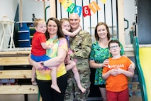 woman in multi-colored tie-dyed t-shirt and black pants holds young boy in red shirt and blue shorts. Man in green camouflage uniform stands in the middle holding a girl in a black tank top. Woman in green, yellow and black shirt holds the shoulders of a boy in an orange t-shirt, shorts and glasses.