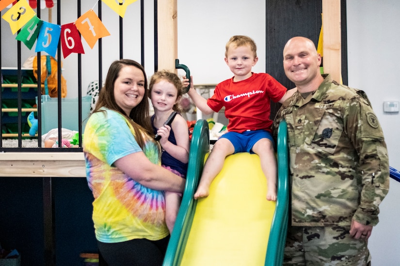 Woman in multi-color tie-dyed shirt holds young girl in black tank top while young boy in red shirt and blue shorts sits at the top of a yellow and green slide and a man in green camouflage uniform stands to the right.