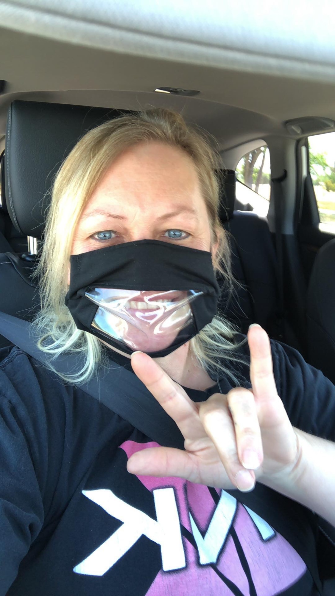 A woman wears a protective mask with a window over the mouth so her lips can be read while using the ALS sign for "I love you."