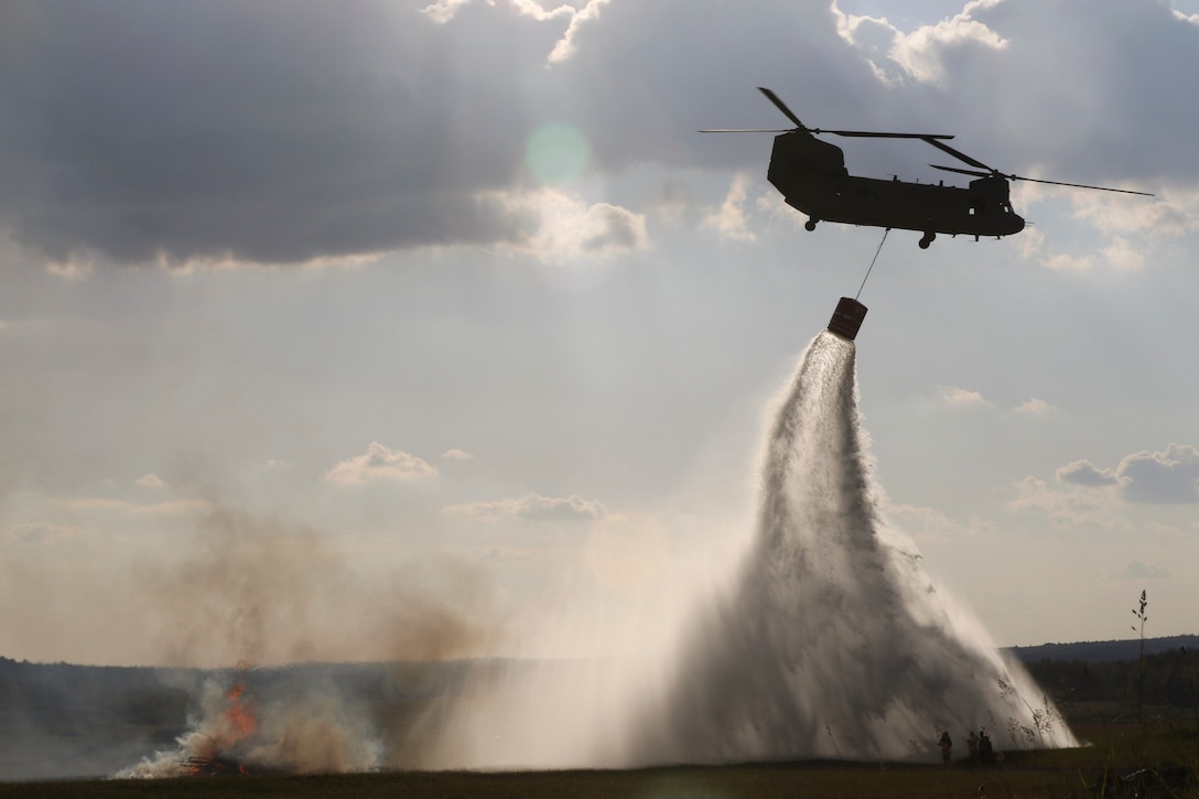 An Army helicopter holds a bucket dropping water by rope.