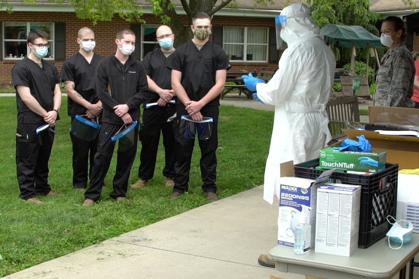 Four men clad in black and wearing face masks listen as a man wearing full personal protective equipment briefs them.